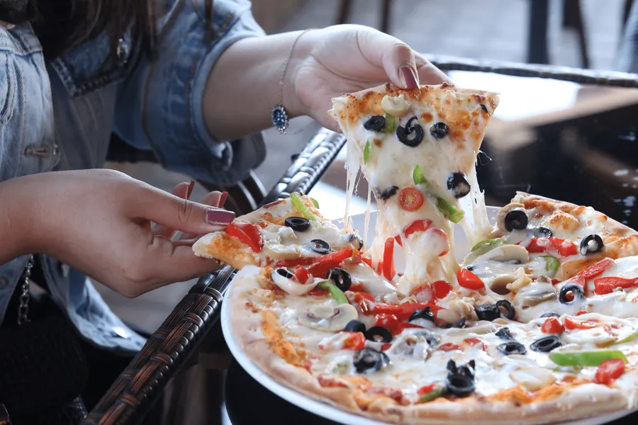 Hands lifting a slice from a vegetable pizza topped with olives and peppers